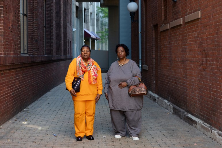 Two women stand in an alleyway flanked by brick buildings. One woman wears a scarf, orange pants and an orange shirt; the other woman wears jewelry, a brown purse and a gray shirt and pants.
