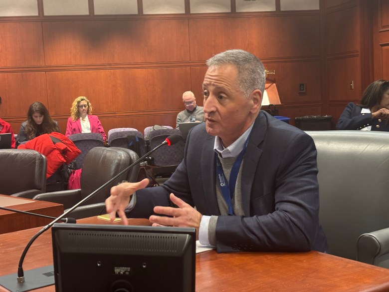 A man sits and speaks at a podium in a legislative committee room.