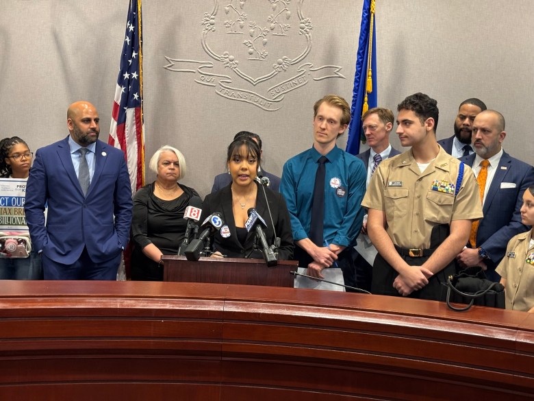 A high school student speaks at a podium with several press microphones attached. Several well-dressed people stand next to her.